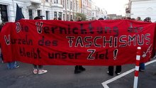Demonstrierende halten hinter ihren Rücken ein rotes Bannermit der Aufschrift "Die Vernichtung der Wurzeln des FASCHISMUS bleibt unser Ziel!"