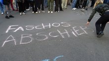 Eine Demonstrierende schreibt mit Kreide auf die Straße "FASCHOS ABSCHIEBEN", im Hintergrund sieht man zahlreiche Demonstrierende stehen.