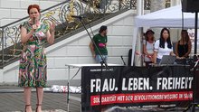 Links im Bild ist Oberbürgermeisterin Katja Dörner zu sehen, die eine Ansprache auf der Kundgebung hält. Rechts davon befindet sich der Stand von FrauLebenFreiheit Bonn mit seinem Banner samt Aufschrift "Solidarität mit der Revolutionsbewergung im Iran", und einige Aktivist*innen, die hinter dem Stand stehen.