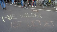 Kreide-Aufschrift "NIE WIEDER IST JETZT!" auf der Straße; im Hintergrund einige Demonstrierende