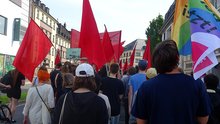 Zu sehen ist ein Demontrationszug durch die Bonner Innenstadt von hinten, mit vielen roten Fahnen und einer bunten von verdi. Rechts und links der Straße ist Bebauung zu sehen.