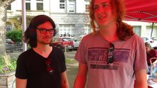 Zu sehen sind Moritz Butscheid (dunkle, längere Haare, Brille, dunkles T-Shirt) und Tristan Kock (blonde, längere Haare, graues T-Shirt mit Aufdruck) in der Nähe des Infostandes der Linken Bonn.