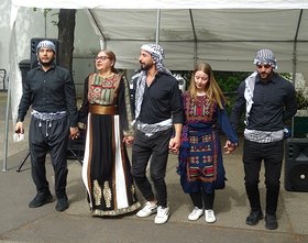 Palästinensische Tanzgruppe; 5 Personen; außen und in der Mitte 3 Männer, schwarz gekleidet mit Palästinensertüchern auf dem Kopf und um die Hüften; dazwischen 2 Frauen mit traditionellen Kleidern