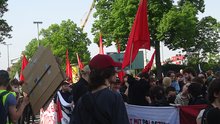 Demozug von leicht seitlich/von hinten; im Vordergrund ist ein junger Mensch mit längerem Haar und rotem Käppi von der Seite zu sehen, sein Gesicht ist aber nicht erkennbar.  Einige Demonstrierende halten Palästina-Banner hoch.  Im Hintergrund sind Bäume zu sehen.