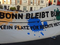 Demonstrierende halten ein großes weißes Transparent, das bunte Farbkleckse aufweist so wie die Botschaft: "BONN BLEIBT BUNT. KEIN PLATZ FÜR RECHTE HETZE."